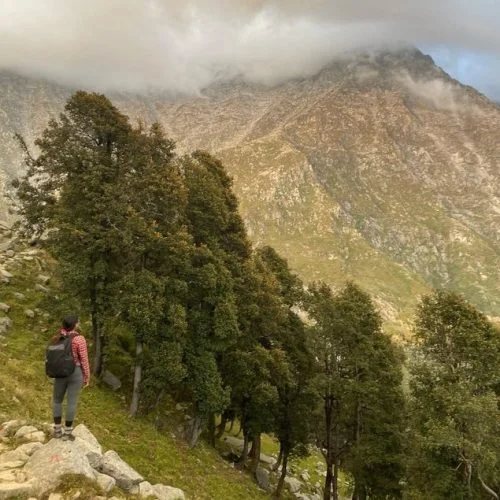 The image shows a female digital nomad in India looking at tall mountains in Dharamkot
